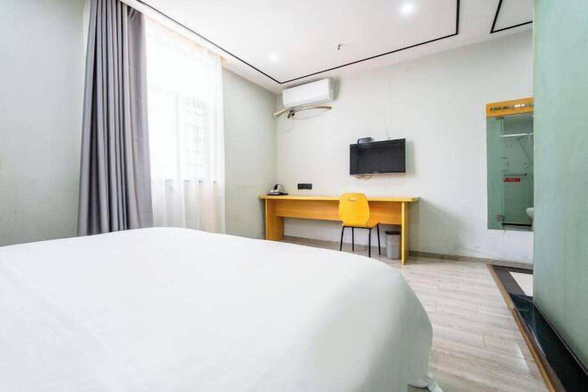 Une chambre d’hôtel comprenant un lit, un bureau, une chaise, un appareil de climatisation, une télévision et une salle de bain avec une porte en verre.