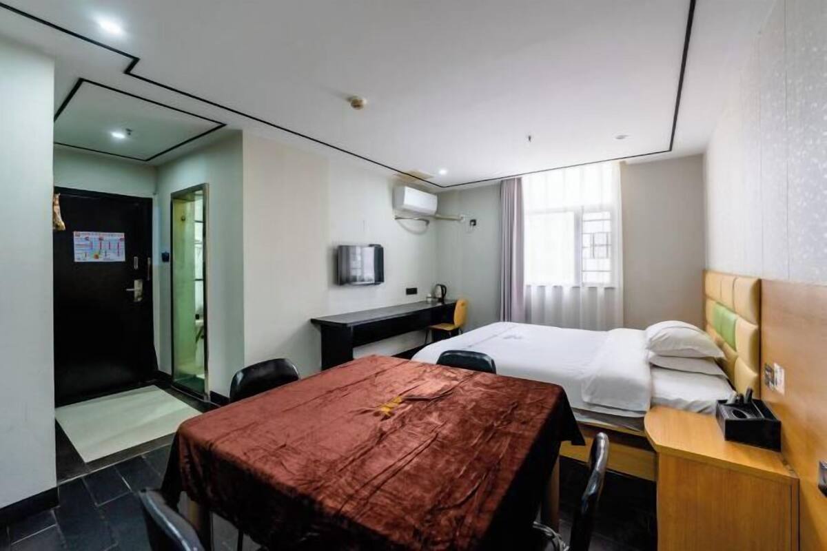 Une chambre d’hôtel avec un lit, un bureau, une chaise, une table recouverte d’une nappe marron, une télévision et une fenêtre avec des rideaux.