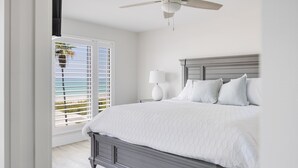 Ein Schlafzimmer mit einem großen Bett, einem Deckenventilator, einem Fenster mit Jalousien und Blick auf den Strand.