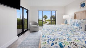 A bedroom with a large bed, a chair, a nightstand, and a view of the outdoors.