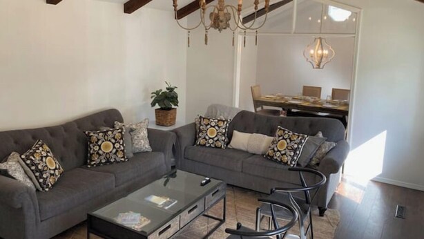 A living room with a dark grey sectional sofa, a glass coffee table, and a chandelier. There is a dining area in the background with a table and chairs. - The Castle at Camorene

 (Springfield)