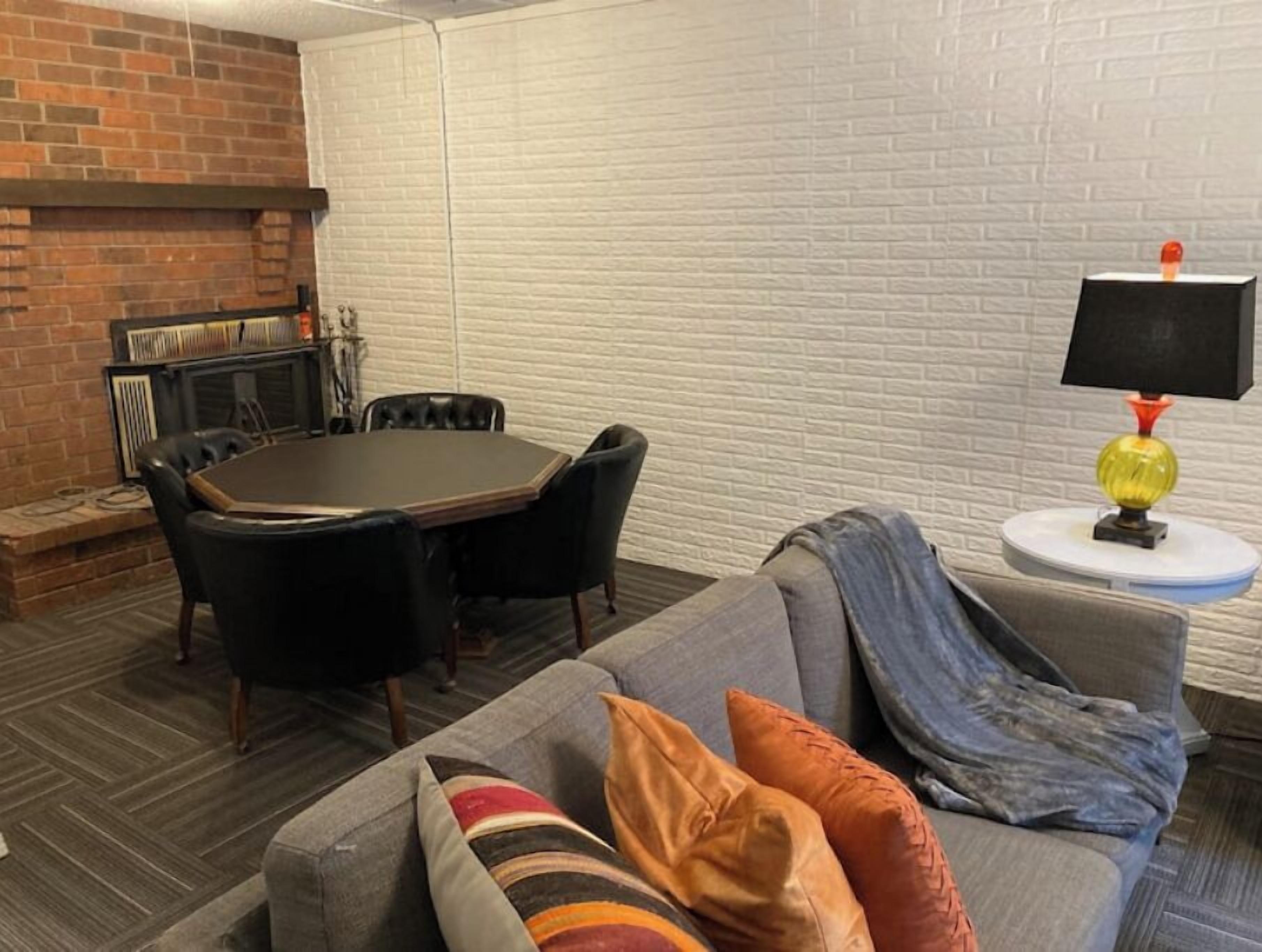 A living room with a brick fireplace, a round table with chairs, a grey sofa with colorful cushions, and a lamp on a side table.