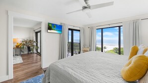 Une chambre avec un grand lit, un ventilateur de plafond et une vue sur l’océan par les fenêtres.