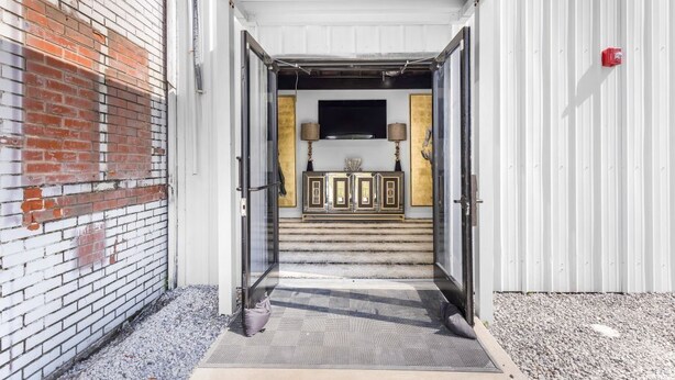 A building entrance with a brick wall, a white door, and a set of stairs leading to a room with a television and a cabinet. - The Sanctuary Spacious 1BR Condo. Stay in Comfort! (Biloxi)