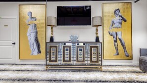 Une pièce dotée d’un tapis à motifs de marbre, d’une console avec un cadre doré et de deux grands tableaux encadrés d’or représentant une figure masculine.