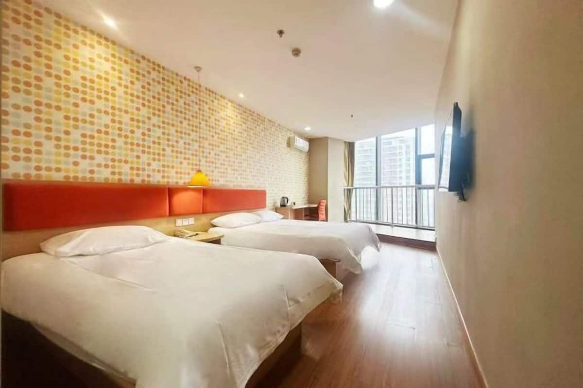 A hotel room with two beds, a red headboard, a yellow lamp, and a large window with a view of buildings.