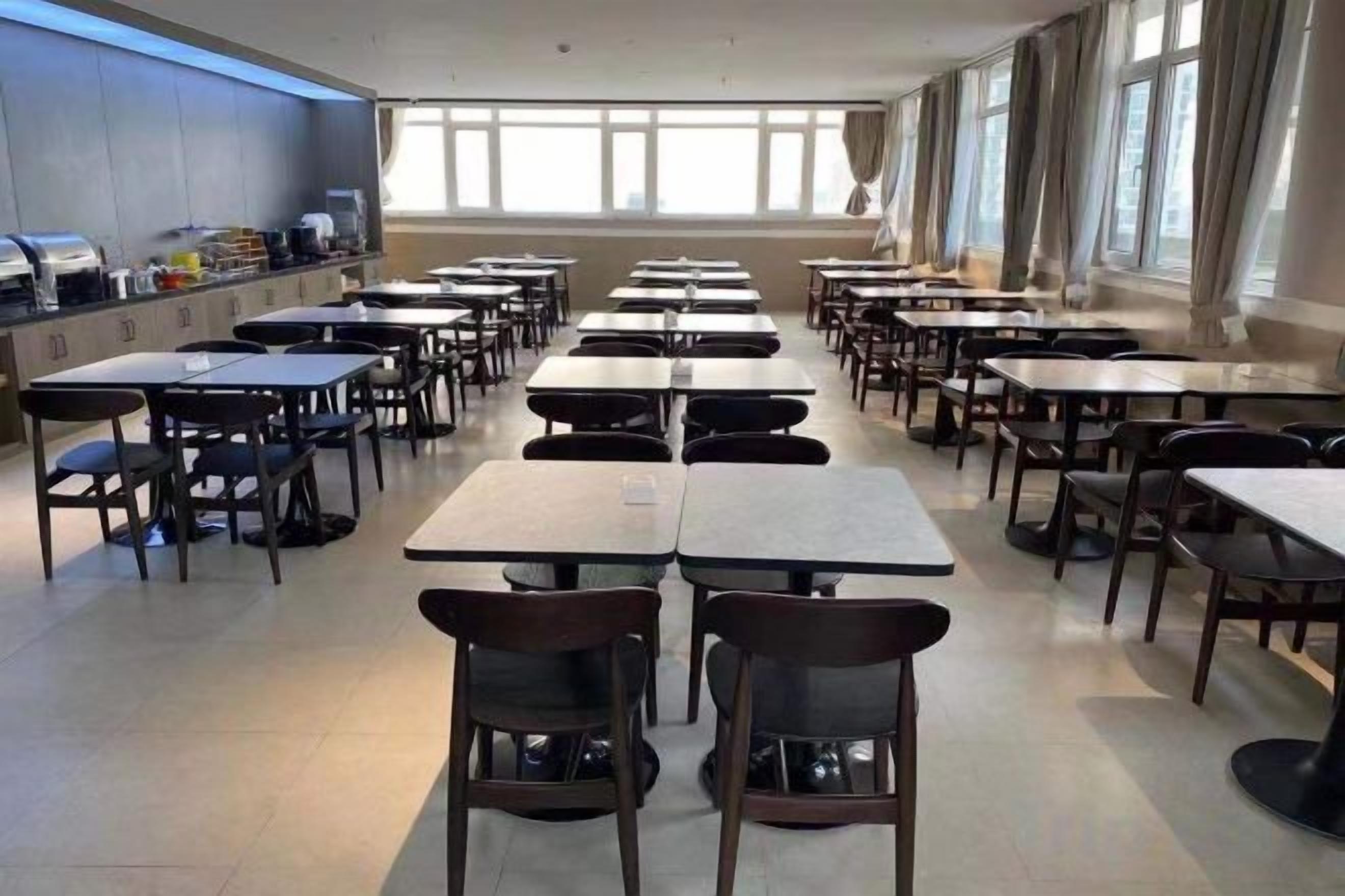 A cafeteria with multiple tables and chairs arranged in rows, large windows on the right side, and a counter with food items on the left.