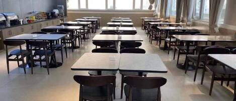 A cafeteria with multiple tables and chairs arranged in rows, large windows on the right side, and a counter with food items on the left.