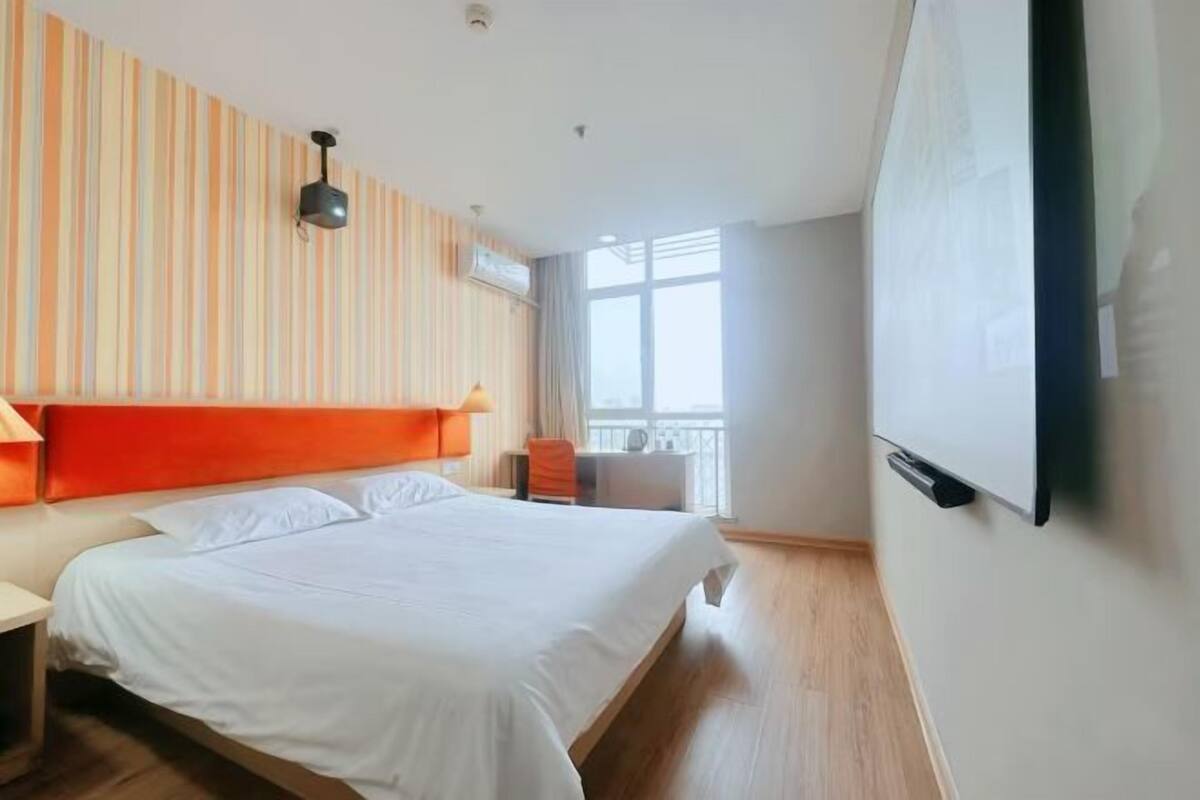A hotel room with a large bed, a TV mounted on the wall, and a window providing natural light.