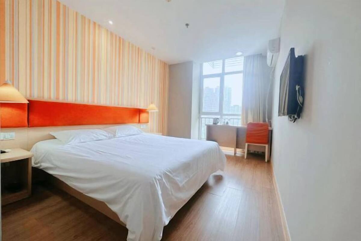 A hotel room with a large bed, a desk, a chair, a TV mounted on the wall, and a window with a view of the city.