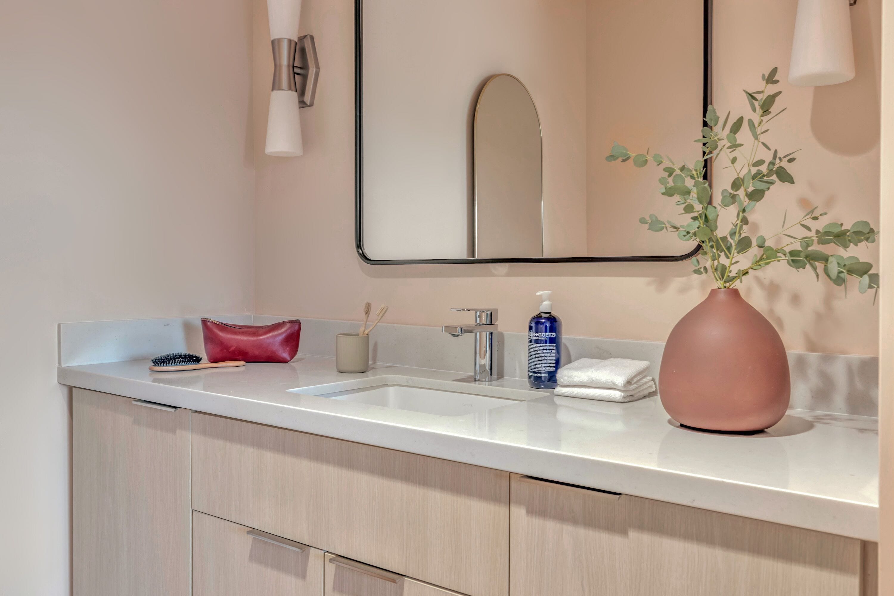Un bagno moderno con un piano d'appoggio bianco, uno specchio, un lavandino, una borsa rossa, una bottiglia blu e un vaso con foglie verdi.