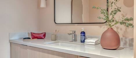 Un bagno moderno con un piano d'appoggio bianco, uno specchio, un lavandino, una borsa rossa, una bottiglia blu e un vaso con foglie verdi.