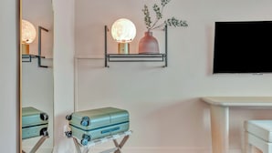 Una habitación con un espejo grande, un televisor en la pared, una mesita pequeña y una maleta sobre un atril.