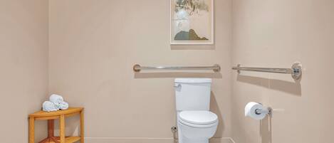 A bathroom with a toilet, a towel rack, a small wooden table with towels, and a framed picture on the wall.