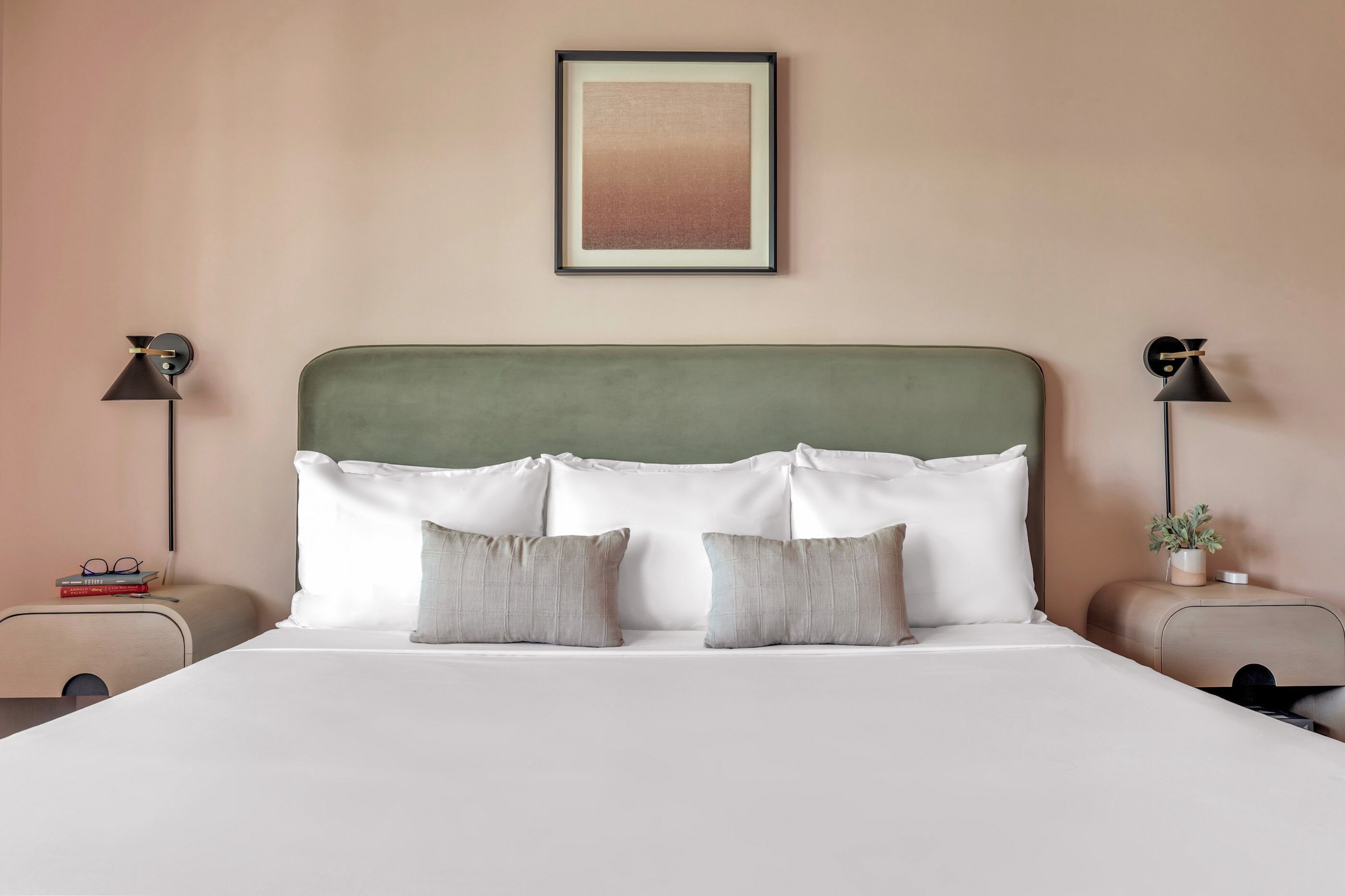 A neatly made bed with white linens and two pillows, flanked by two bedside tables with lamps and a small plant.