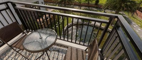 Un balcon avec une petite table et deux chaises, une rambarde en métal, et une vue sur un jardin et un élément décoratif aquatique.