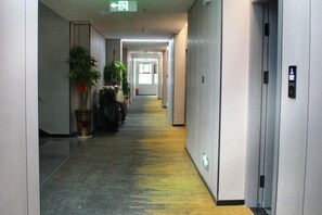 A long, narrow hallway with a yellow carpet, a trash bin, and a potted plant on the left. - Sweet Orange Hotel (Hengyang)
