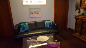 A living room with a black leather sofa, a glass coffee table with purple legs, and a sign above reading "PENSION RIVERSIDE".