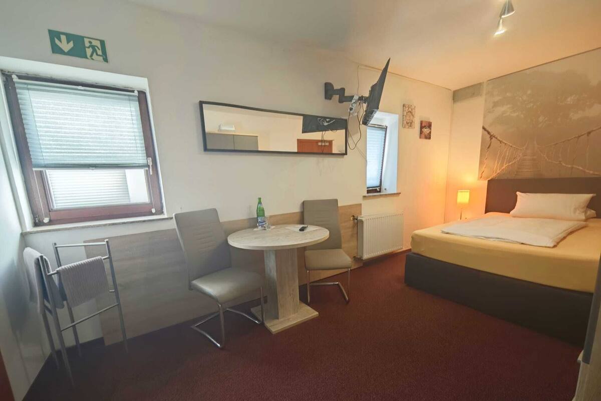 A compact hotel room with a bed, a small round table with a chair, a radiator, and a window with blinds.