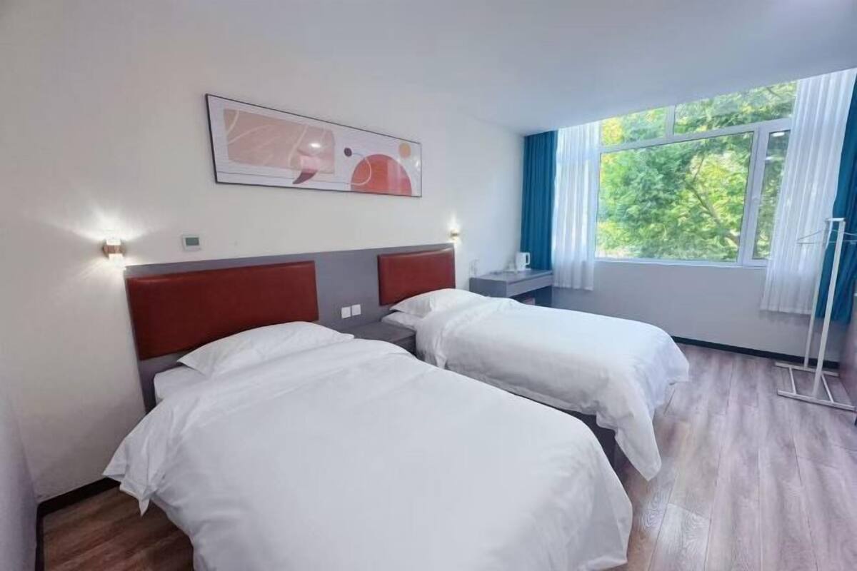 Quarto de hotel com duas camas, uma janela com cortinas, um quadro na parede e uma mesinha de cabeceira.