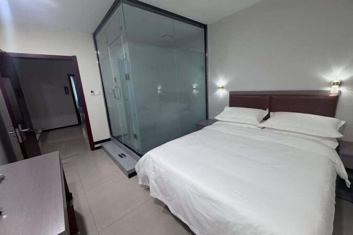 Quarto de hotel com cama, box de vidro, mesa de cabeceira de madeira e luminária fixada na parede.