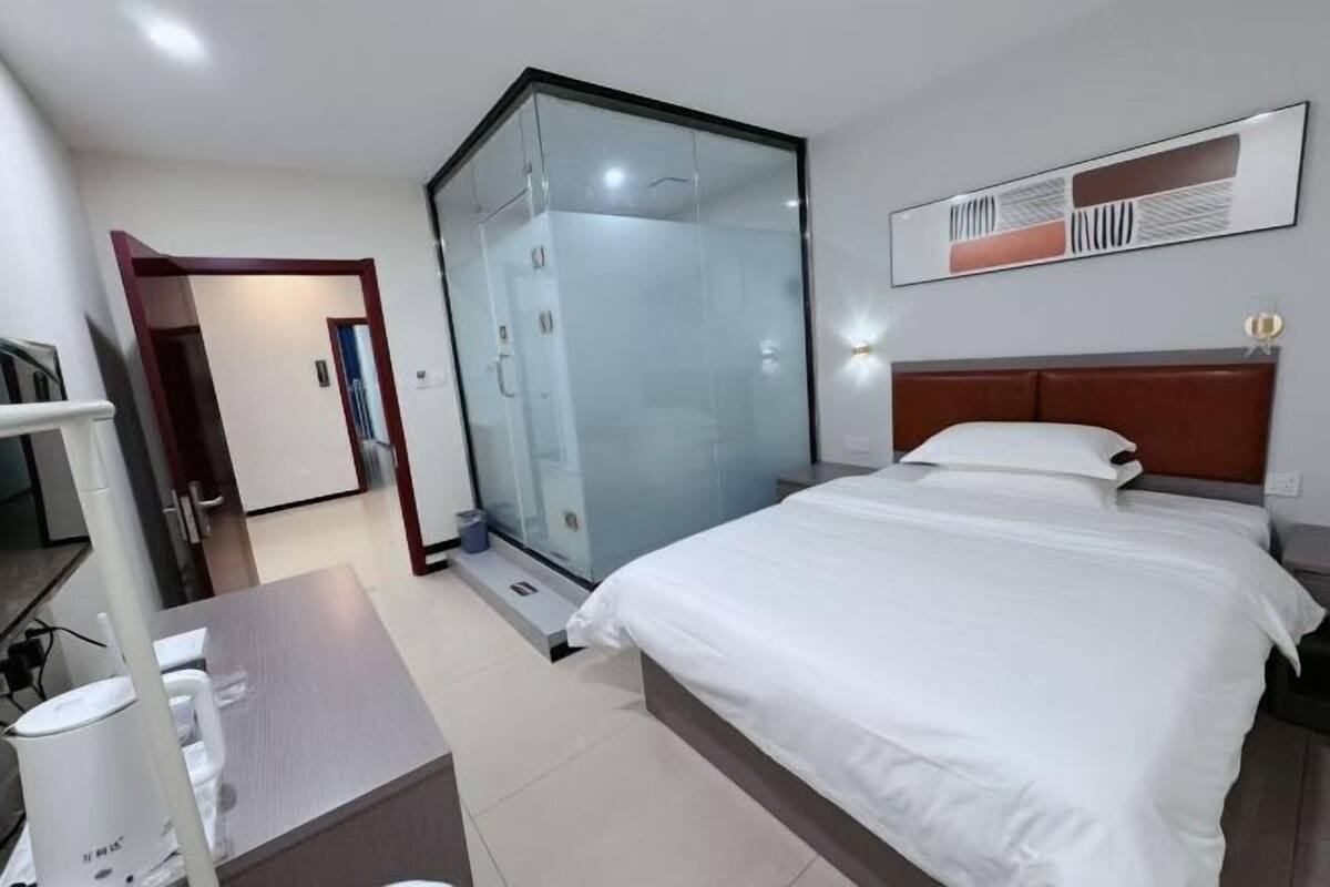 Quarto de hotel com uma cama, uma escrivaninha, chuveiro em gabinete de vidro e luminária fixada na parede.