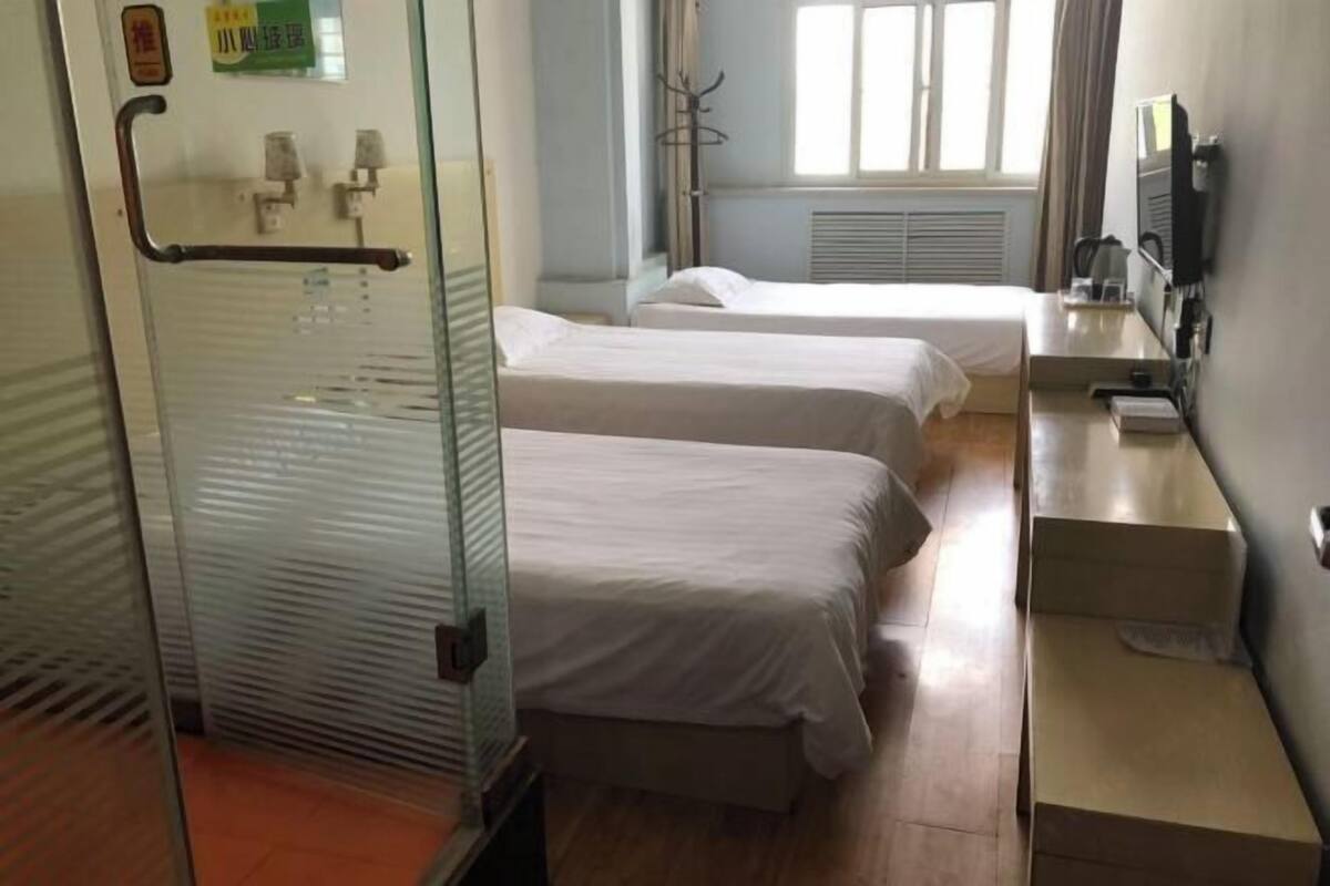 Quarto de hotel com duas camas de solteiro, divisória de vidro, pequena escrivaninha com cadeira e janela com cortinas.
