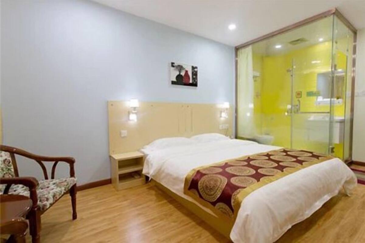 Quarto de hotel com cama, uma cadeira, mesa de cabeceira e box de chuveiro de vidro.
