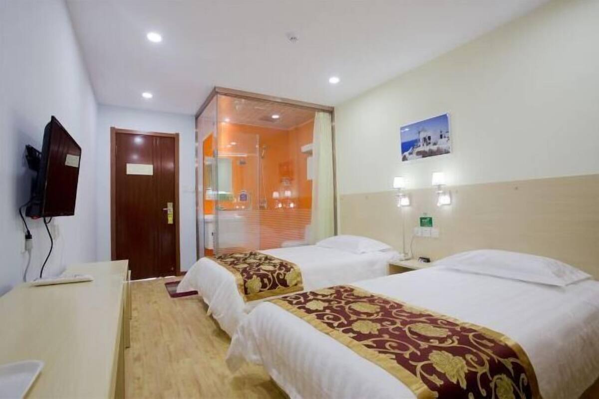 Quarto de hotel com duas camas, uma escrivaninha, televisão e banheiro visível por uma porta de vidro.