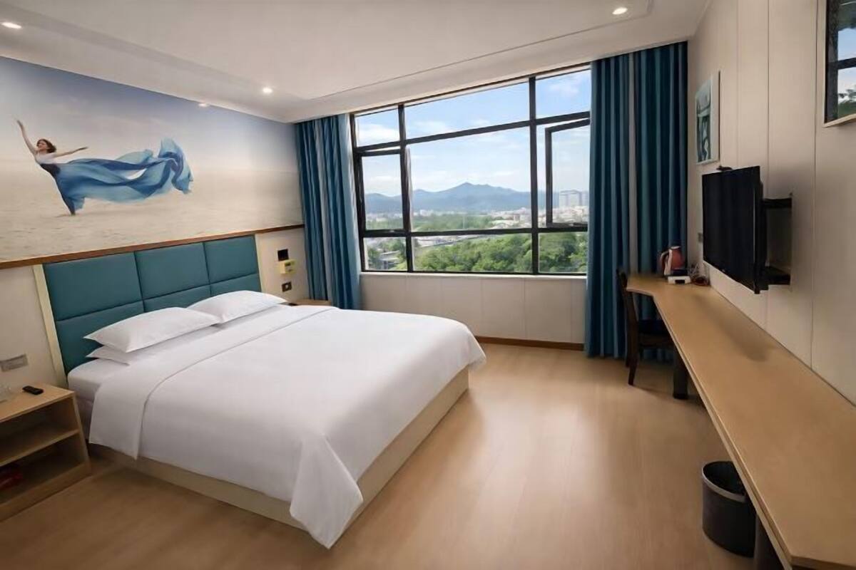 A hotel room with a bed, a desk, a TV, a window with a view of greenery and mountains, and a mural on the wall.