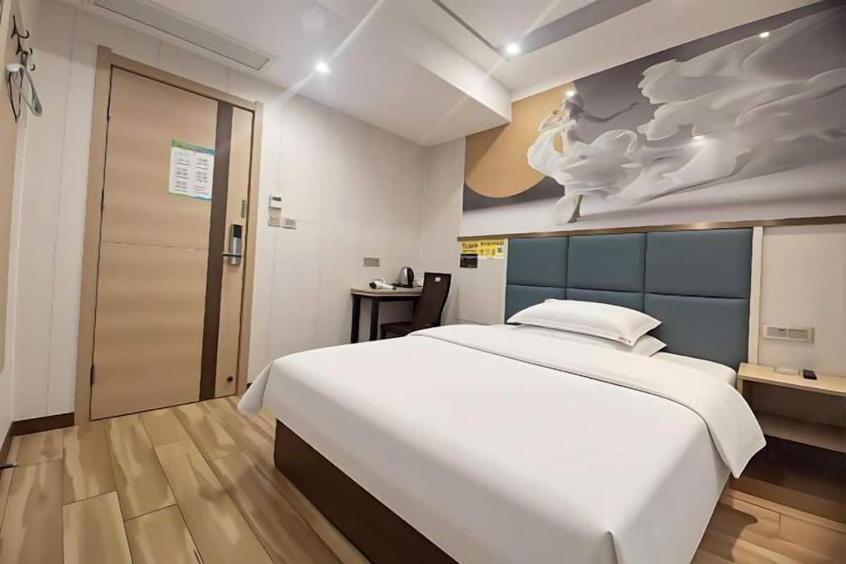 A hotel room with a bed, a desk, a chair, a wall-mounted clock, and a door.