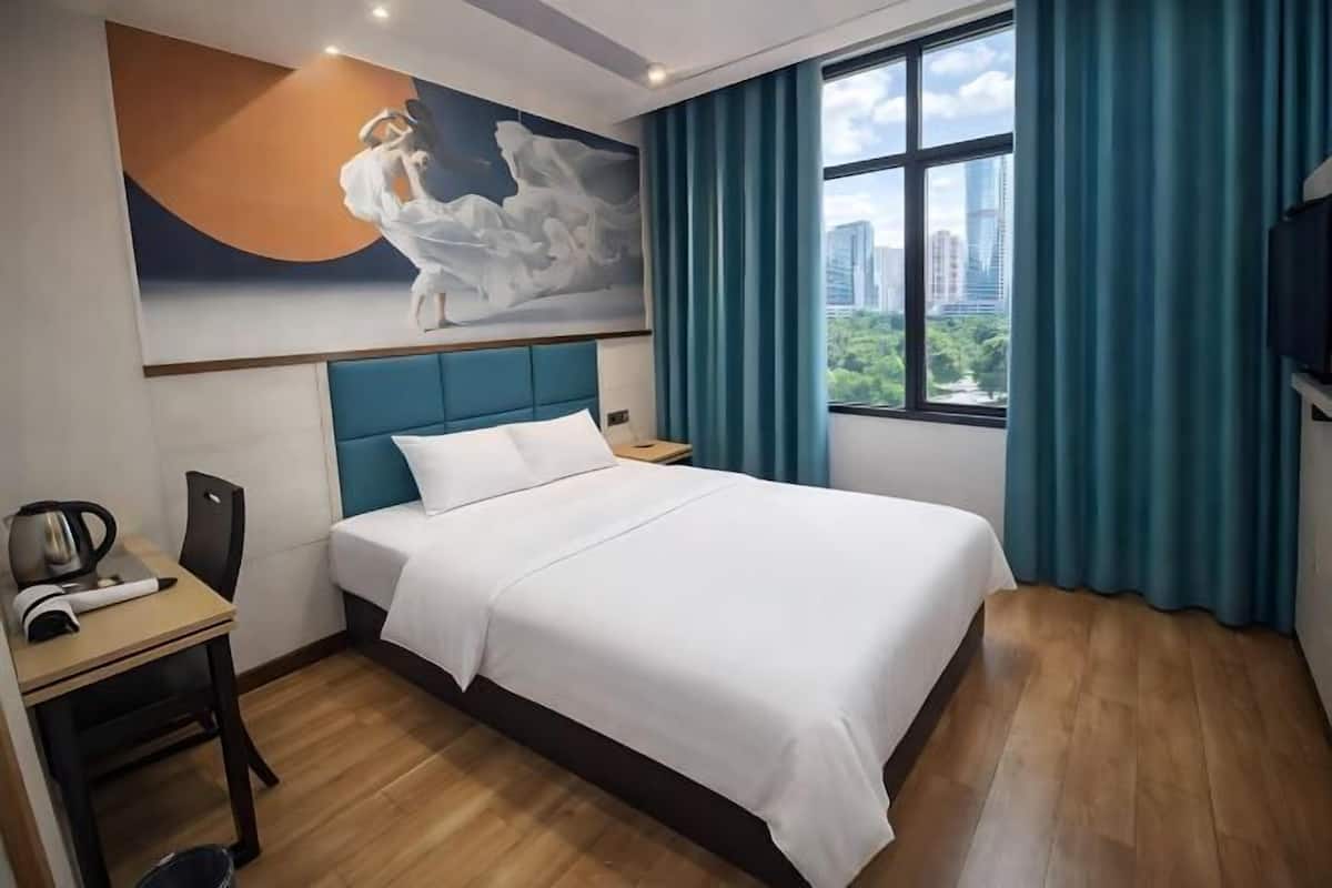 A hotel room with a bed, a desk with a kettle, a chair, a painting on the wall, and a window with curtains.