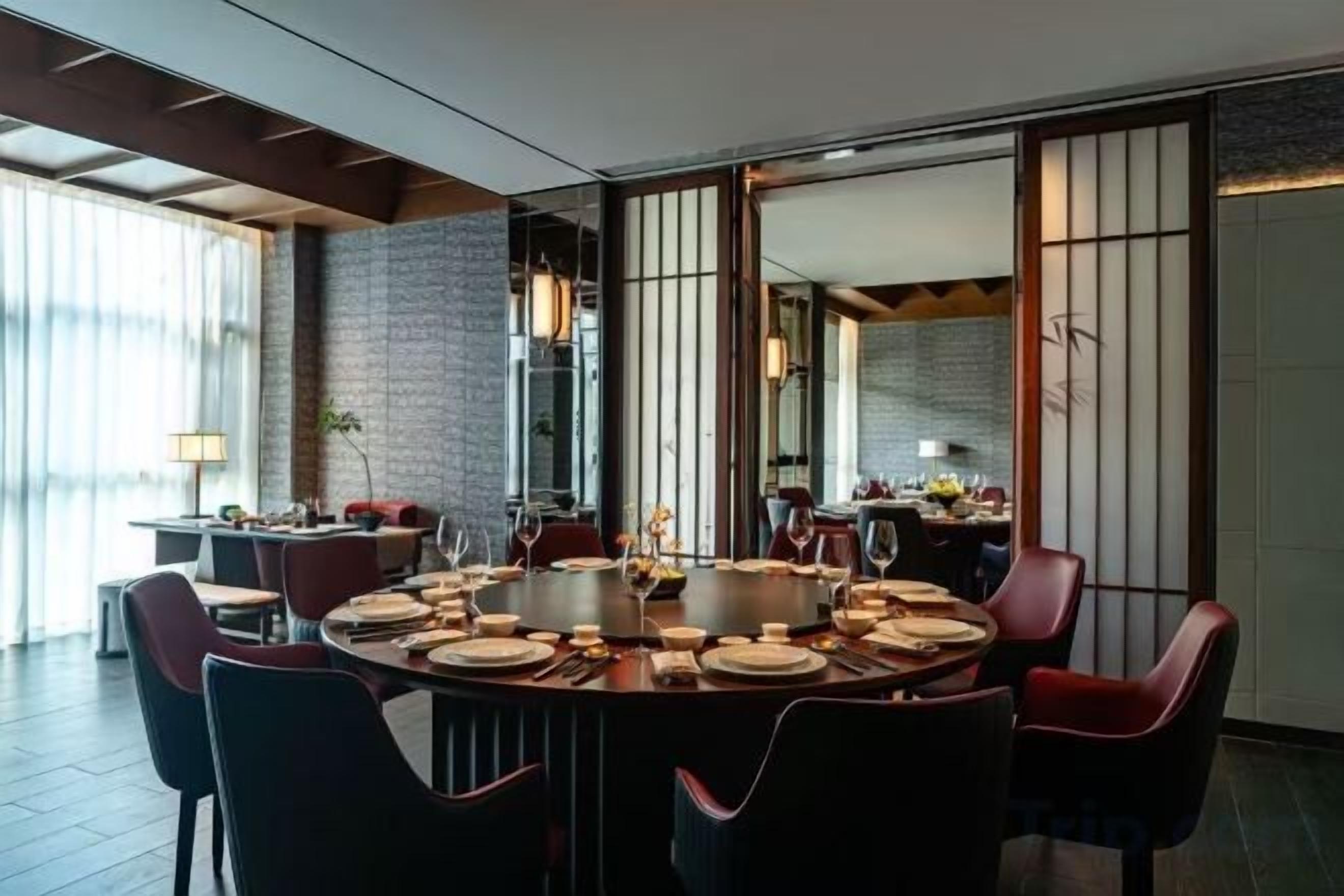 A dining area with a round table set for six, surrounded by chairs, in a modern interior with a brick wall and large windows.