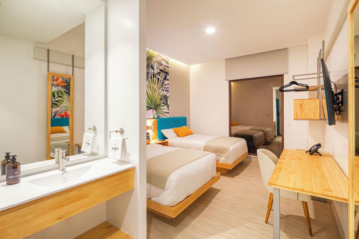 Une chambre d’hôtel moderne avec un grand miroir, une coiffeuse en bois, deux lits avec des oreillers orange, un bureau et une grande plante.