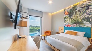A modern hotel room with a large bed, a desk, a chair, a TV mounted on the wall, and a large window offering a view of the outdoors.