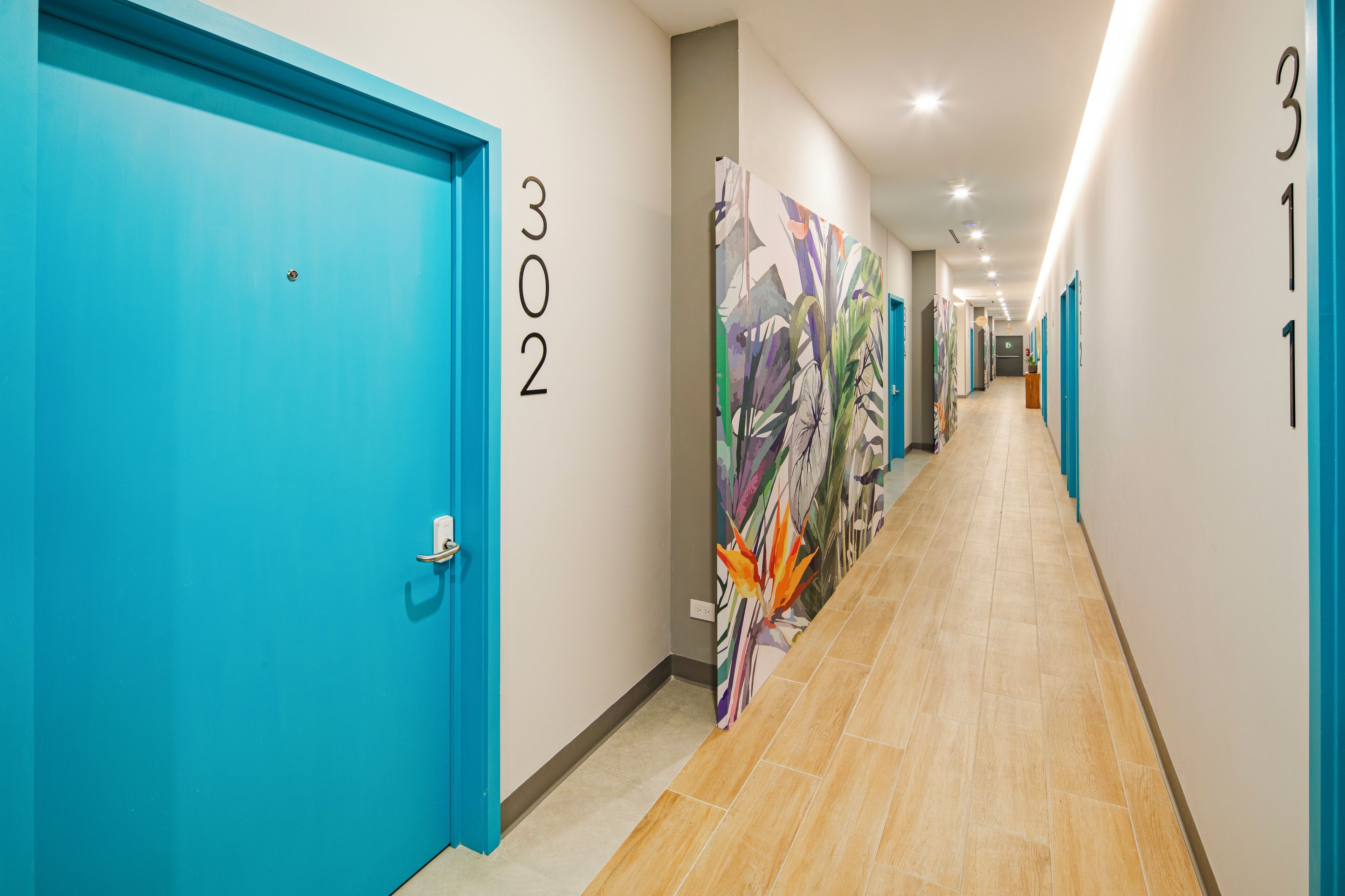 En hotels korridor med turkise døre nummereret 302 og 311, samt et farverigt abstrakt mural på væggen.