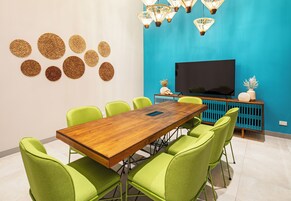 Un salón de conferencias con una mesa de madera y sillas verdes, un televisor de pantalla plana grande y decoración en las paredes.