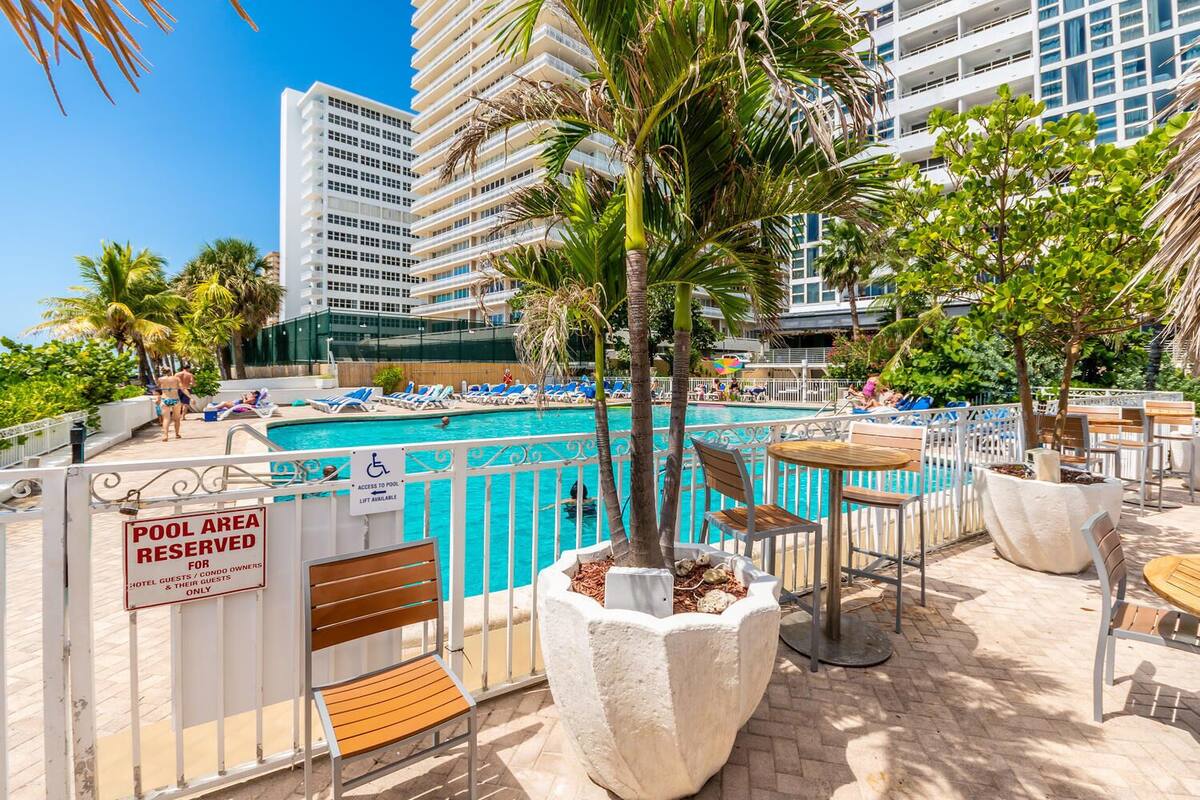 A pool area with a sign indicating it is reserved for hotel guests and condo owners. There are lounge chairs and tables around the pool, and a palm tree in the center.