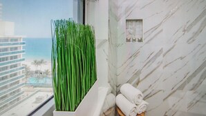 Un bagno con vista sull'oceano, una parete in marmo e una finestra con piante verdi.