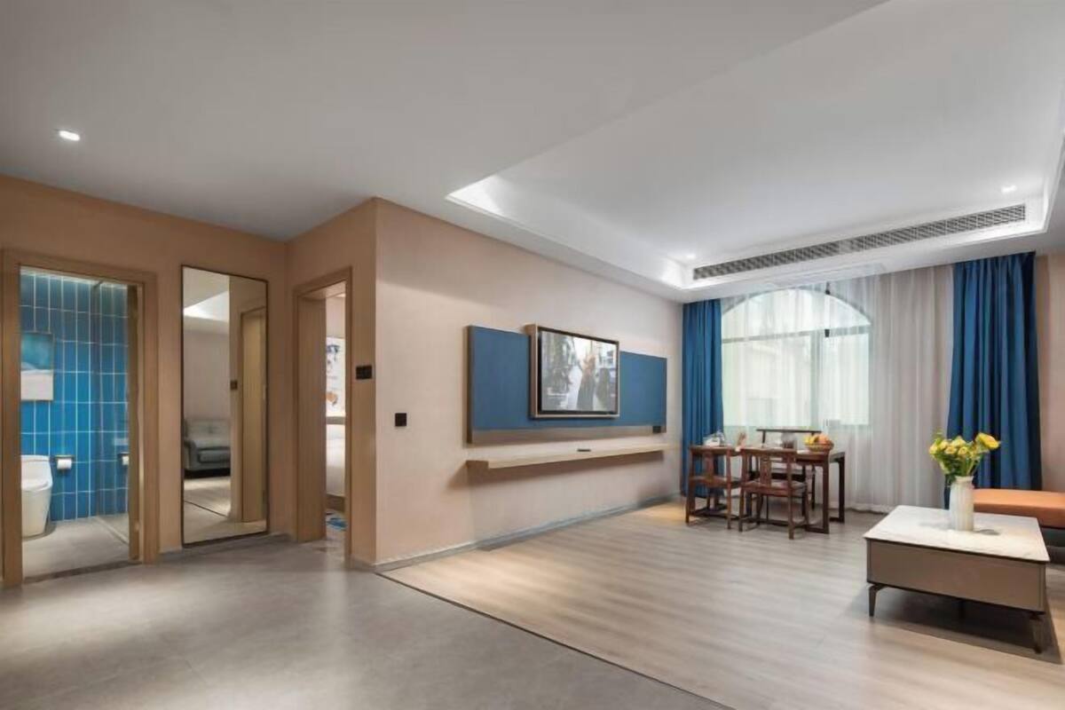 Moderni hotellihuone, jossa on puulattia, ruokailutila pöytineen ja tuoleineen, suuri ikkuna verhoineen sekä seinälle ripustettu taulu.