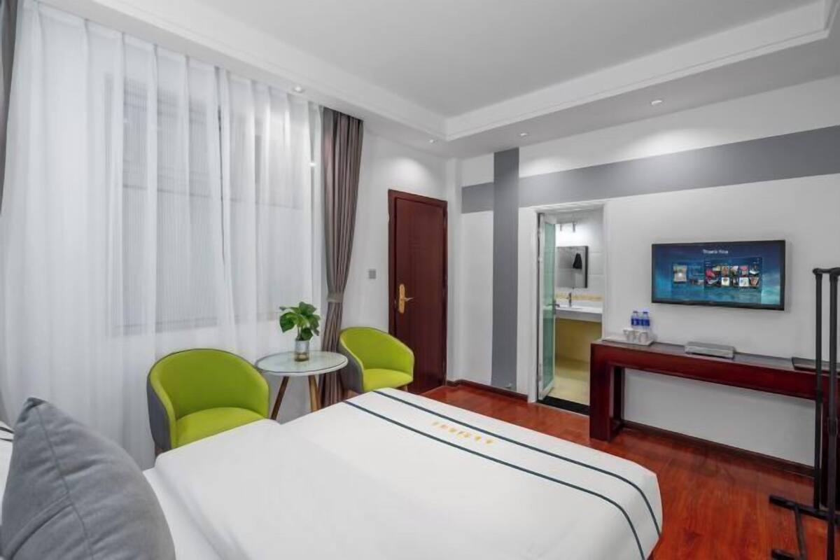 Une chambre d’hôtel avec un lit, deux chaises, une petite table avec une plante, un téléviseur fixé au mur et une porte donnant sur une autre pièce.