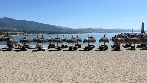 Una spiaggia con file di lettini e ombrelloni di paglia, un cielo azzurro limpido e una montagna sullo sfondo.