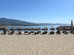 Een strand met rijen ligbedden en rieten parasols, een helderblauwe lucht en een berg op de achtergrond.