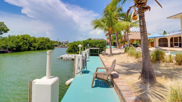 A waterfront dock with a chair, palm trees, and a building in the background. - Walk to Beach: Waterfront Home in Marathon! (Marathon)