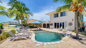 A pool area with lounge chairs, a thatched-roof structure, and a building with a visible sign. - Walk to Beach: Waterfront Home in Marathon! (Marathon)