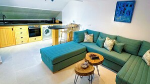 Una sala de estar moderna con un sofá seccional verde, una mesa de centro de madera y una zona de cocina con armarios de madera y una lavadora.