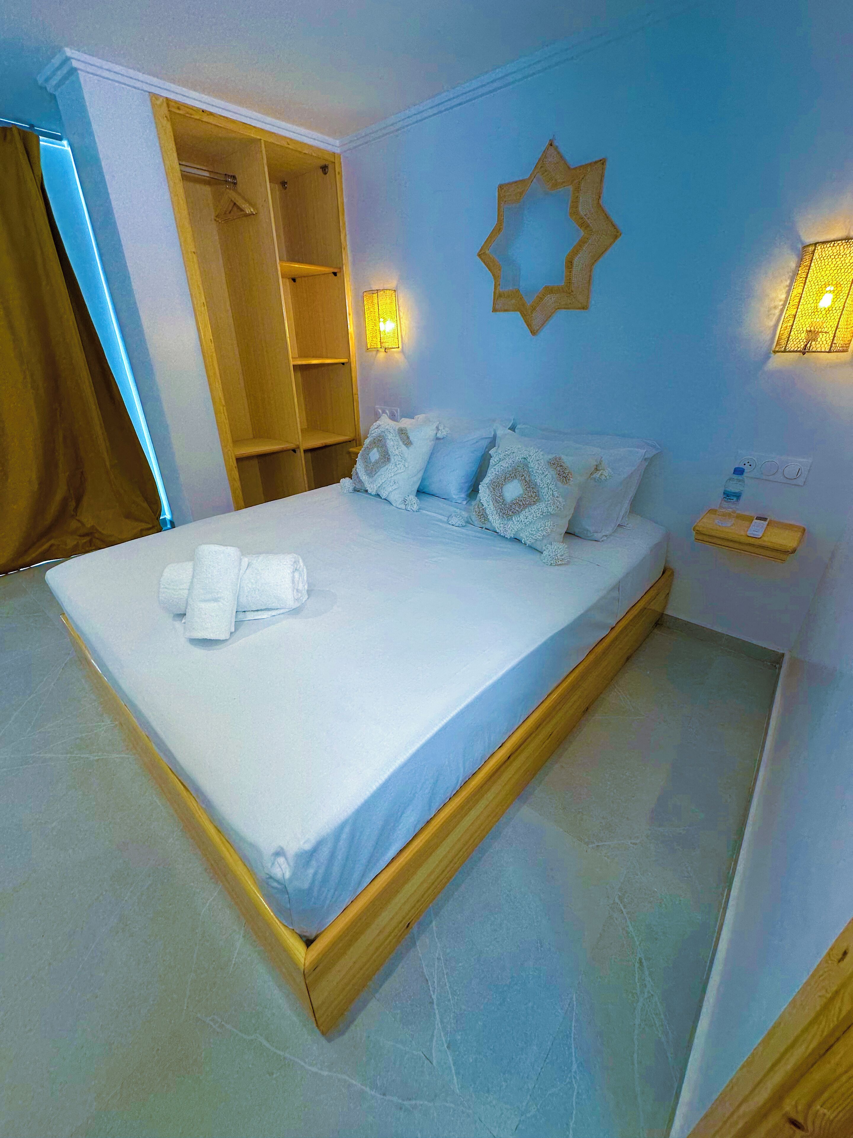 A bedroom with a bed, towels, a nightstand, a wall-mounted light, and a wooden wardrobe.