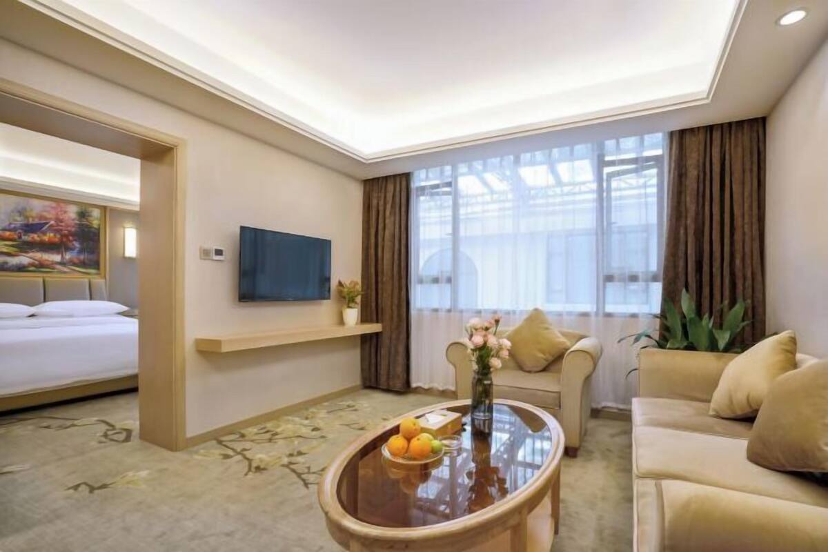 A hotel room with a bed, a sofa, a coffee table with fruit, a TV, and a large window with curtains.