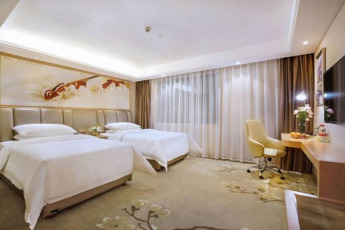 A hotel room with two beds, a desk, a chair, and a large window with curtains.