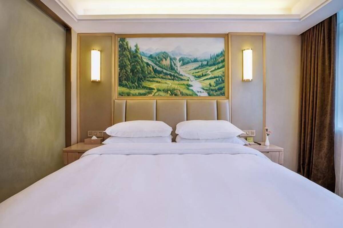 A hotel room with a large bed, two bedside tables, a painting of a landscape, and a window with curtains.
